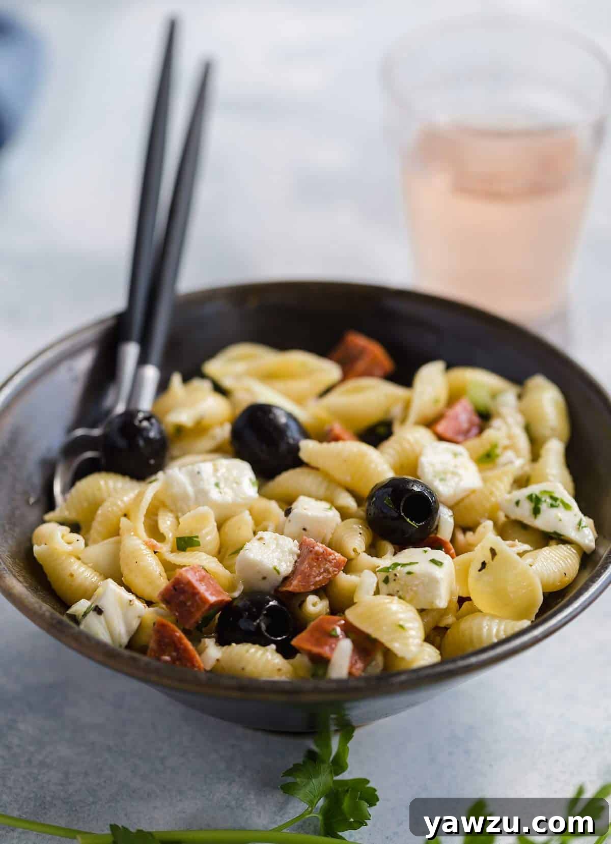 A bowl of vibrant Italian pasta salad with two wooden serving spoons, and a partially obscured glass of wine in the soft-focus background, suggesting a delightful meal.