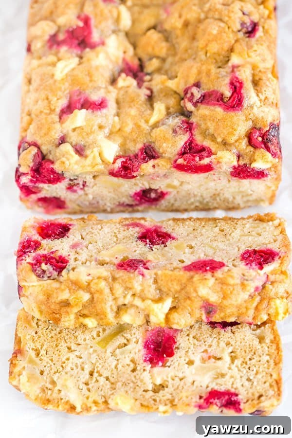 A slice of incredibly moist Apple Cranberry Bread, packed with chunks of apple and bright red cranberries, resting on a wooden board.