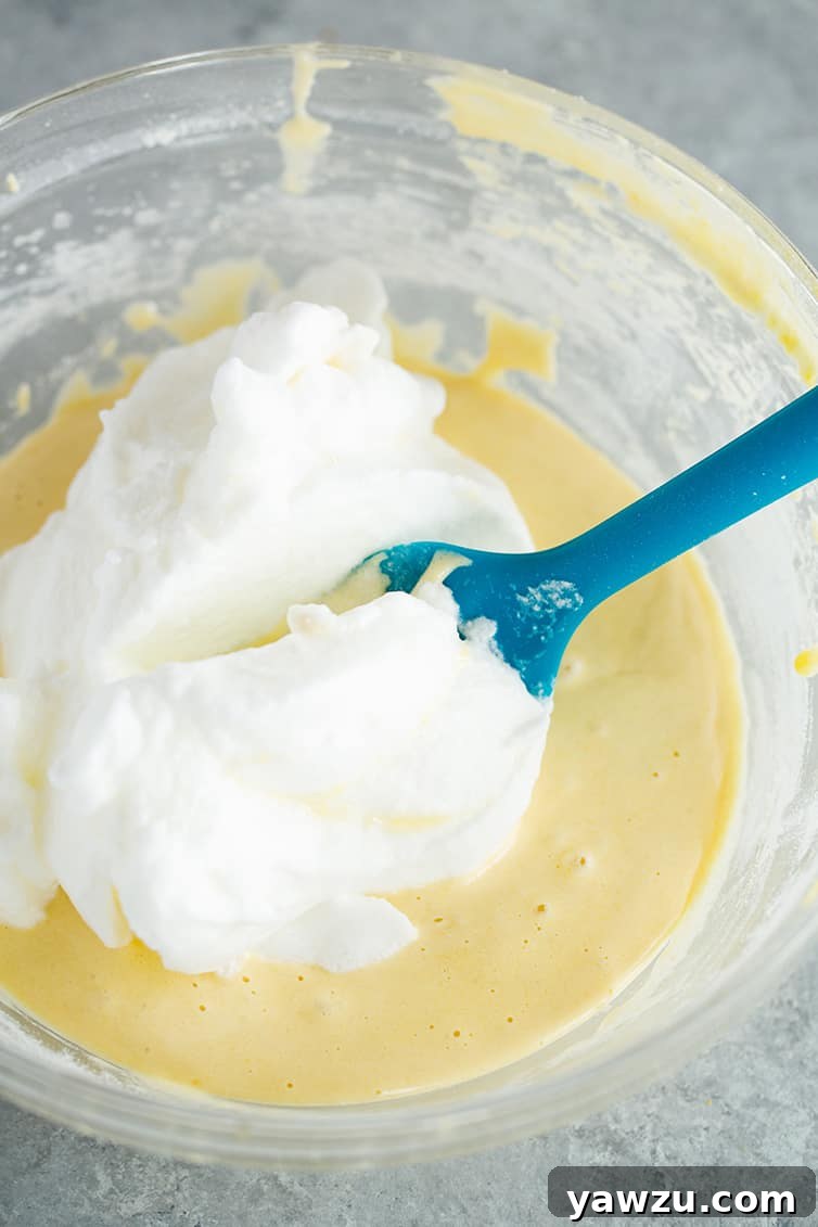 A close-up shot of whipped egg whites being gently folded into yellow cake batter, illustrating the technique for a light cake.