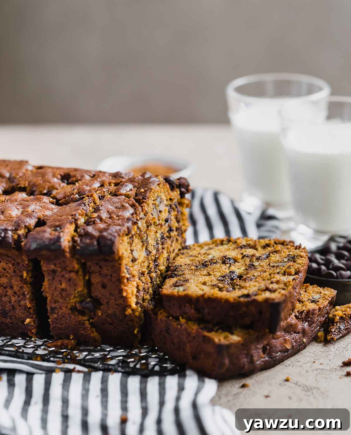Sliced pumpkin bread with chocolate chips and pecans on a tea towel, with glasses of milk in the background. Highlighting its moist texture and rich ingredients.