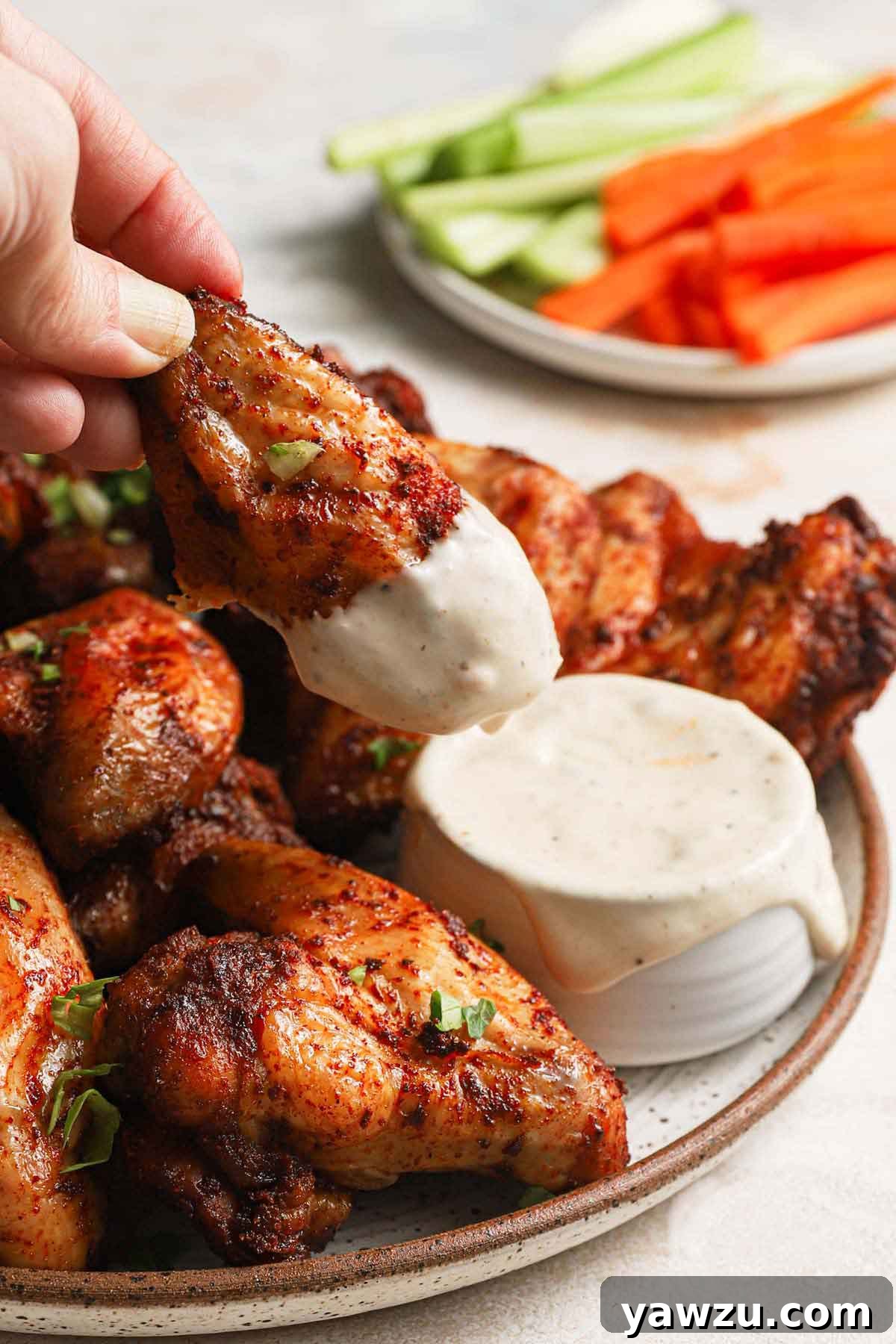 Platter of golden brown, crispy baked chicken wings with one being dipped into creamy ranch dressing. A side plate holds fresh carrot and celery sticks.