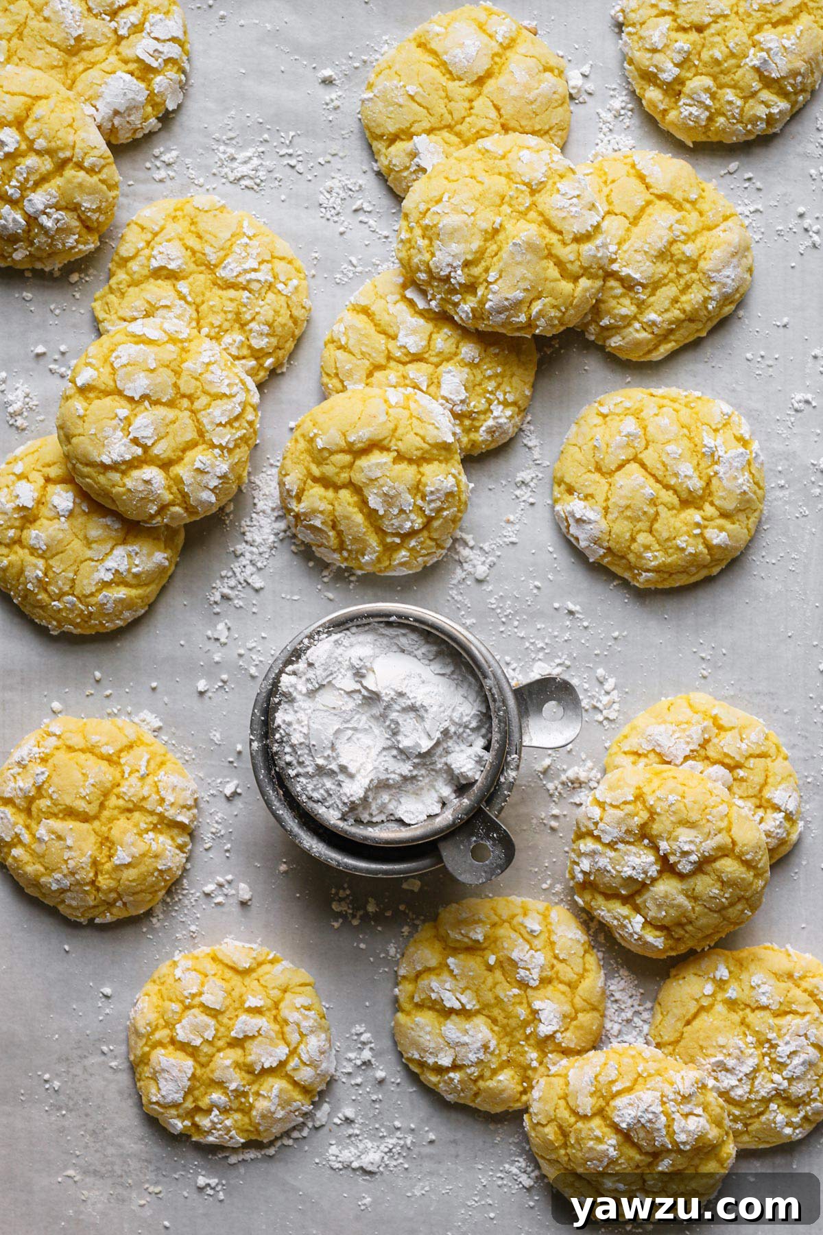 A few lemon crinkle cookies resting on a lined baking sheet dusted in powdered sugar, with a small bowl of powdered sugar in the center.