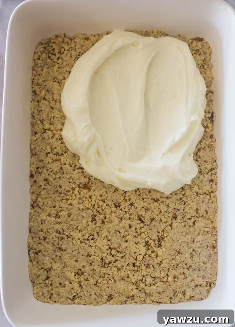 A creamy, sweetened cream cheese layer being smoothly spread over the baked pecan crust.