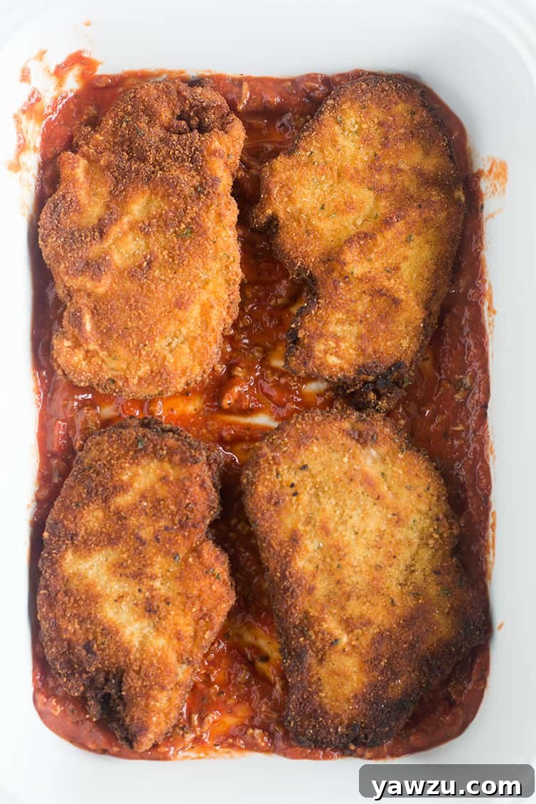 Four golden-brown chicken cutlets resting on a thin layer of rich marinara sauce in a baking pan, ready for cheese.