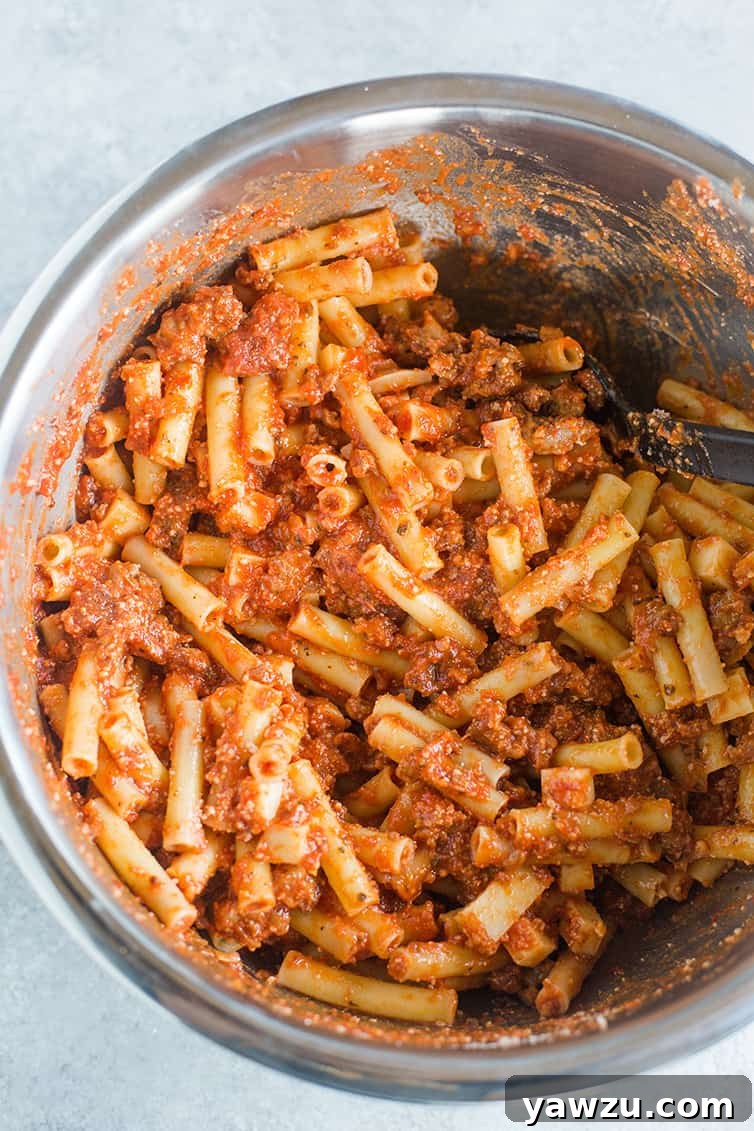 Close-up of baked ziti ingredients mixed together in a large bowl, showing pasta, sausage, and cubed mozzarella coated in sauce, ready for baking.