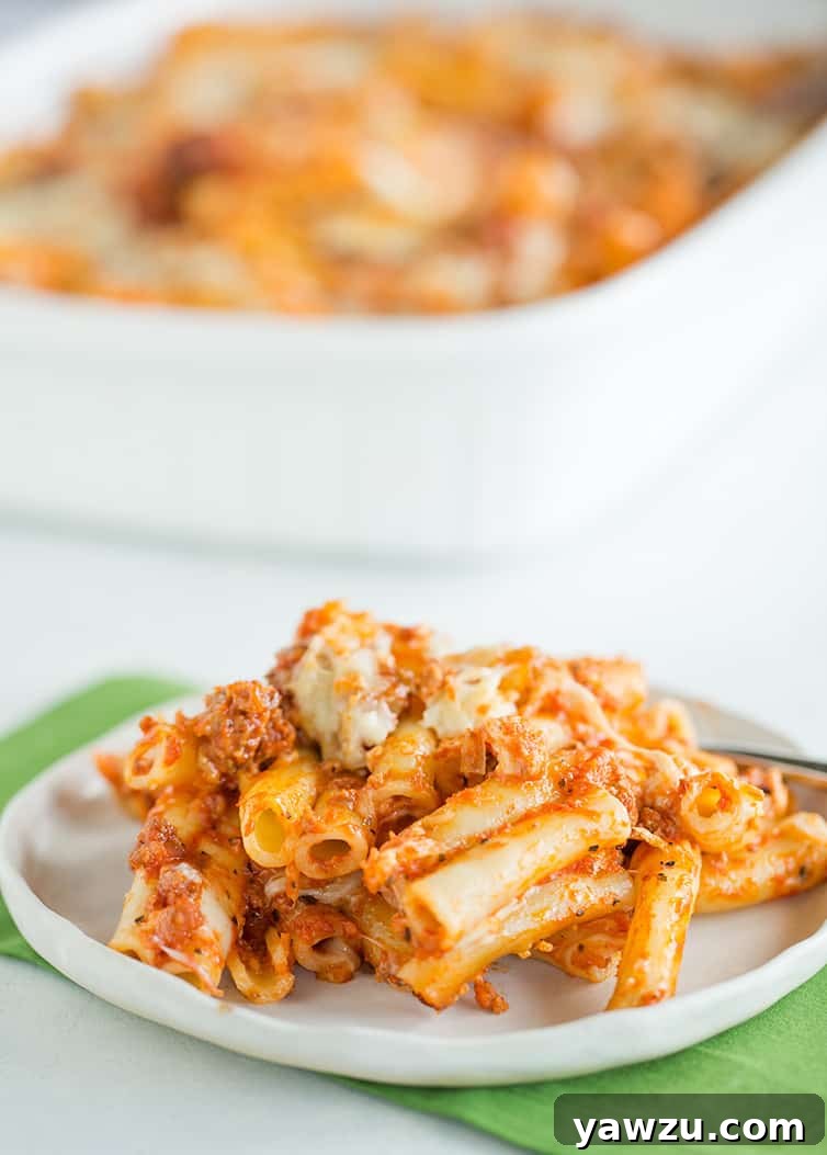 A close-up shot of a plate of baked ziti, showing rich red sauce, melted cheese, and pasta, with the full casserole dish in the background.