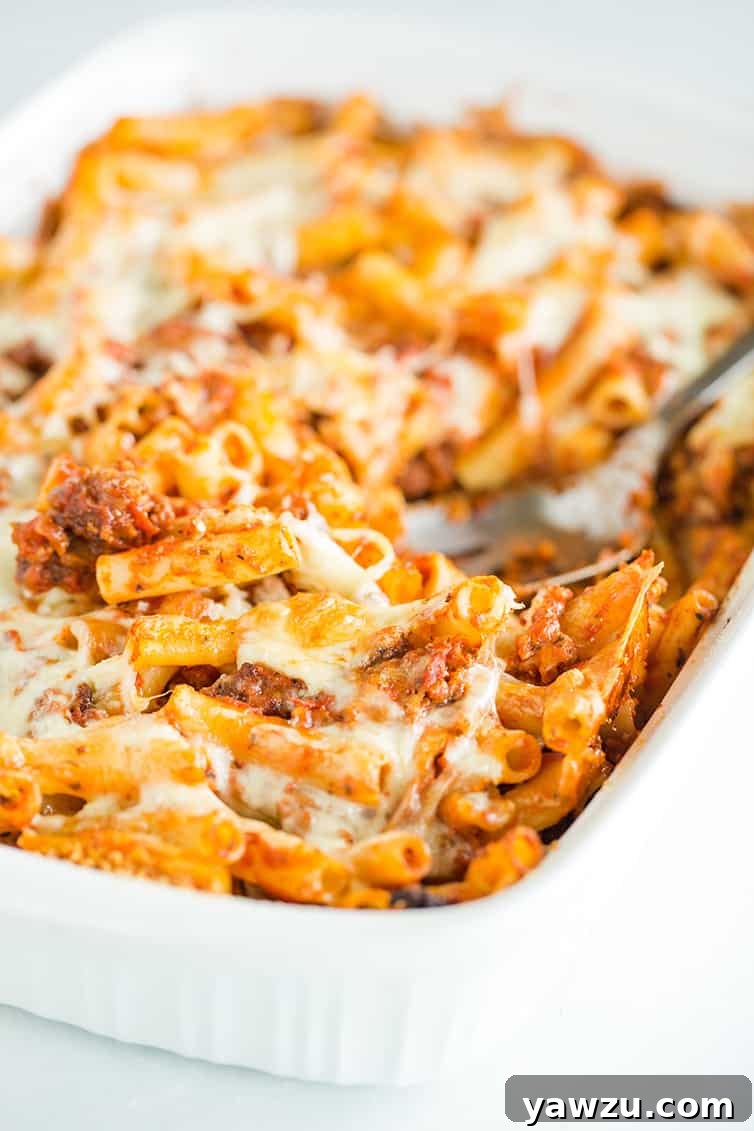 A close-up of a serving spoon lifting a spoonful of baked ziti, revealing layers of pasta, sausage, and melted cheese, from the pan.