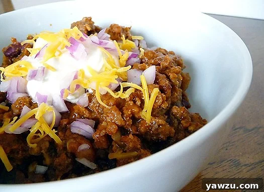 A bowl of rich, hearty chili, generously topped with cool sour cream, finely chopped green onions, and shredded cheddar cheese.
