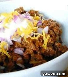 Chili in a bowl topped with sour cream, shredded cheese, and diced onion.