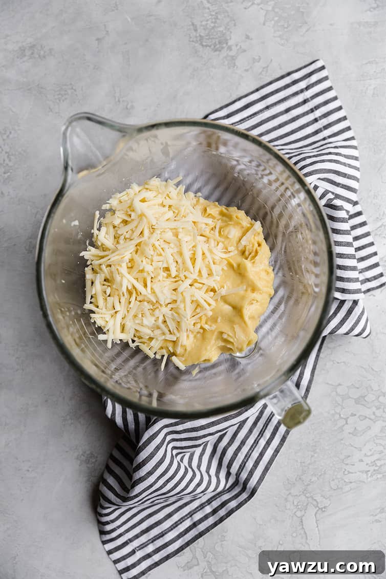 Grated Gruyère cheese being added to the warm pâte à choux dough in a mixing bowl, ready to be incorporated.