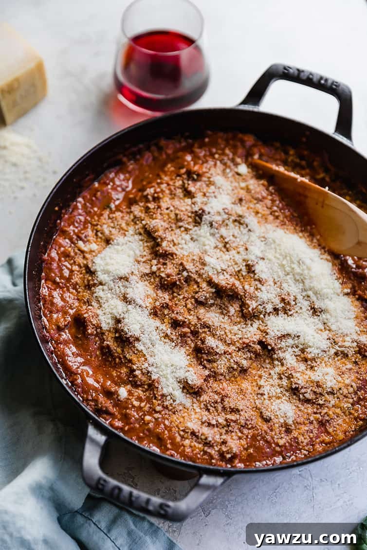 A pan of bolognese sauce being stirred, with grated Parmesan cheese visibly incorporated.