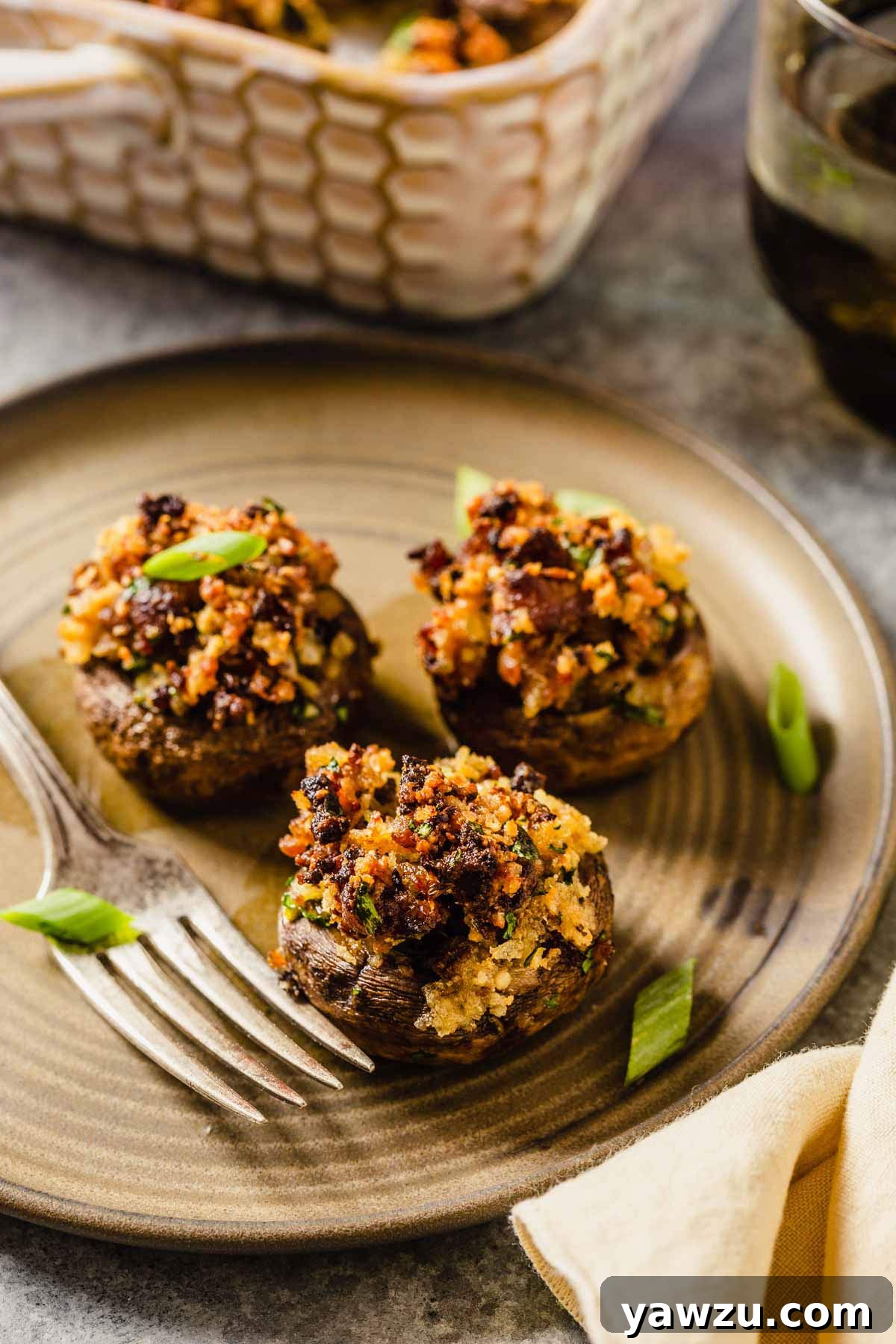 A small white plate featuring three perfectly cooked sausage stuffed mushrooms, garnished with fresh green onion, and a silver fork resting beside them.