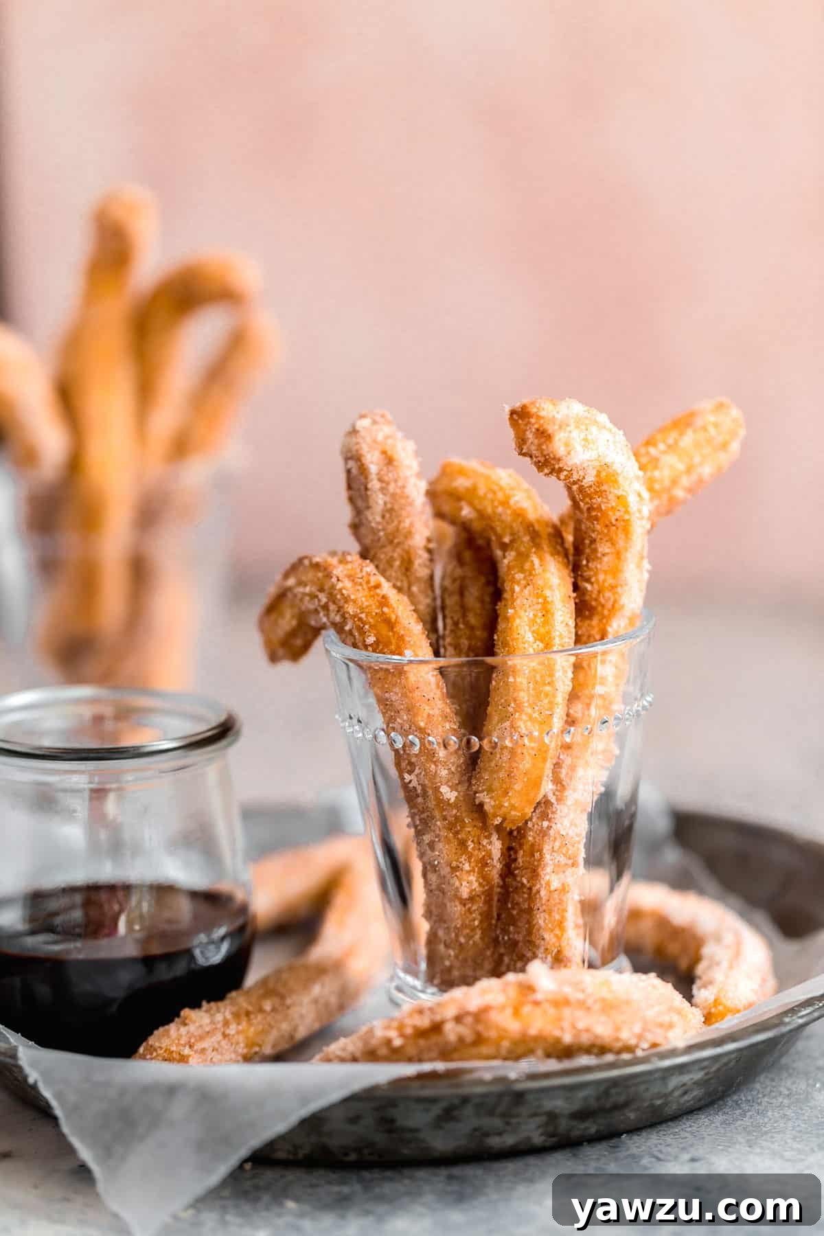 A pie pan with parchment paper with churros in a glass jar on the left and chocolate dipping sauce on the right.