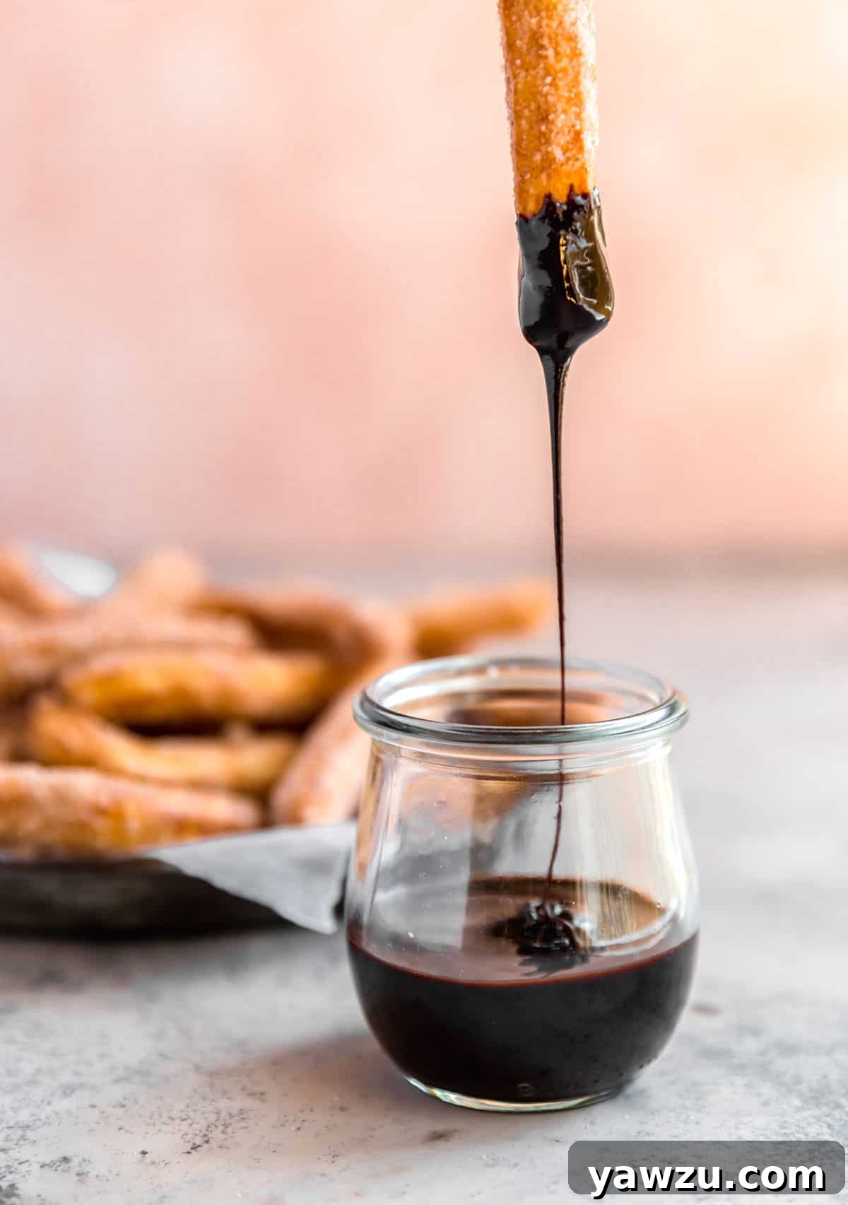 A glass jar with melted chocolate and a churro dipped in with chocolate drizzling back into the jar and a platter of churros behind the chocolate jar.