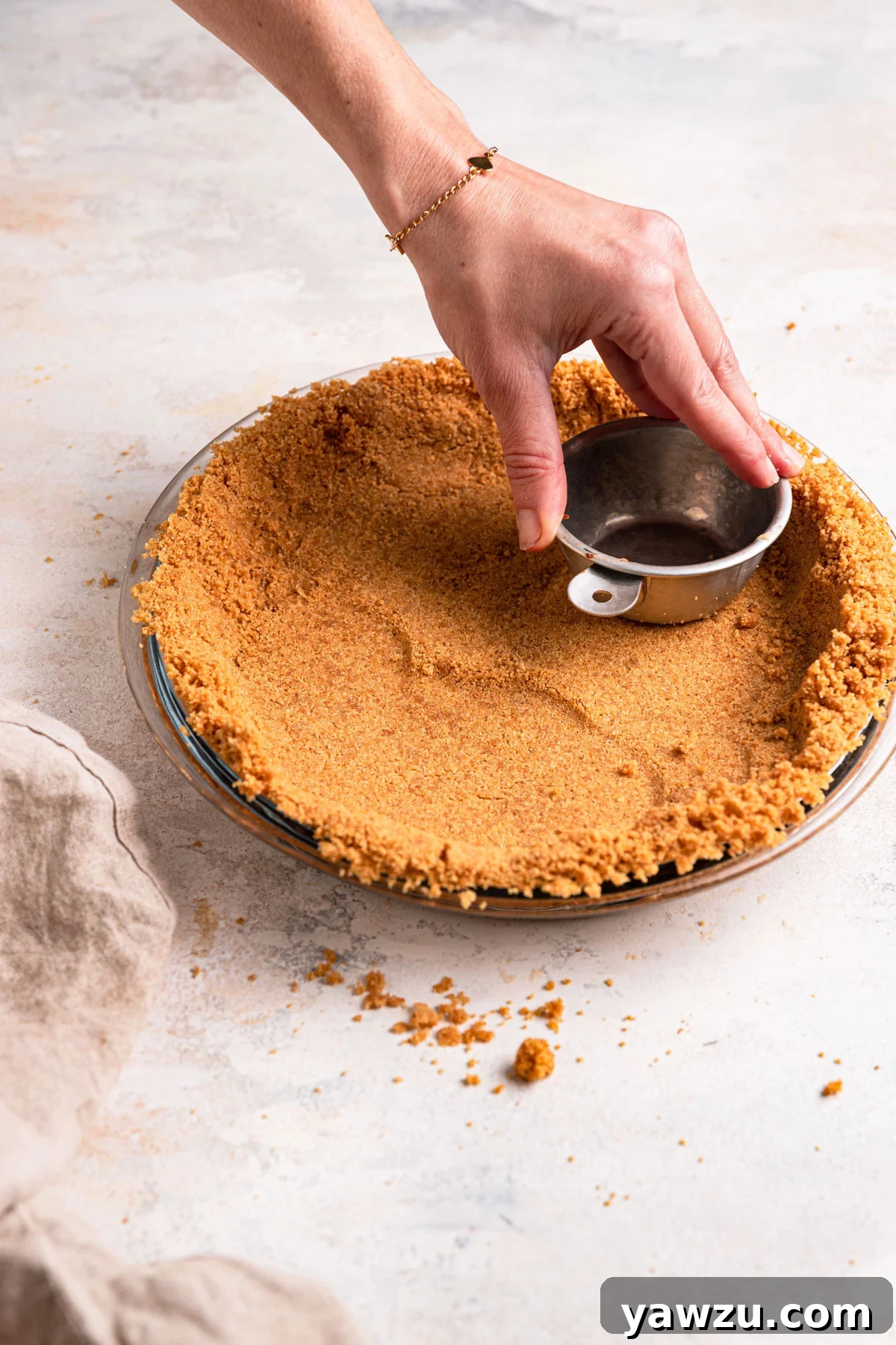 Graham cracker crust being packed into a pie plate using a measuring cup.