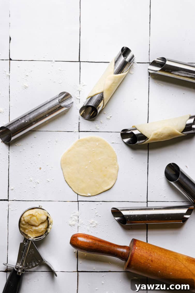 A tile counter with cannoli shell forms a rolling pin a round of dough and a small cookie scoop of dough in the bottom left.