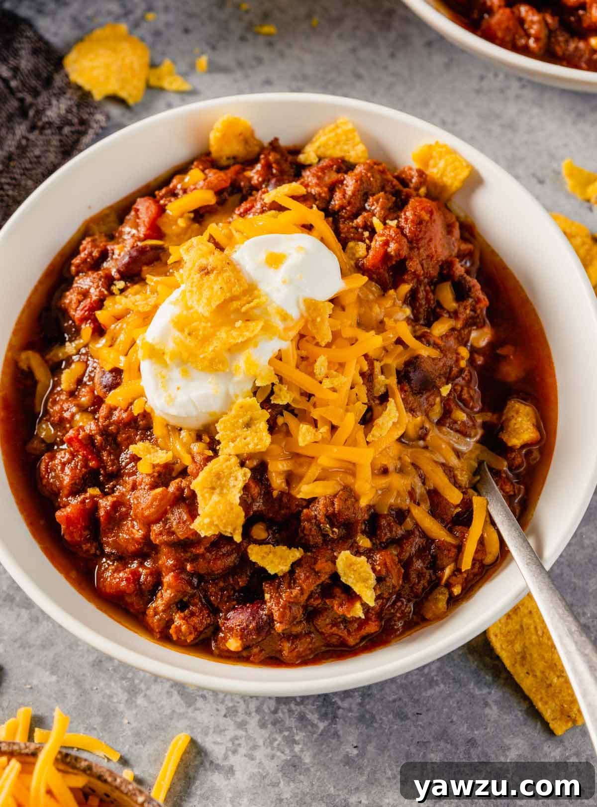 A white bowl brimming with hearty ground beef chili, generously topped with shredded cheddar cheese, crunchy corn chips, and a dollop of sour cream.