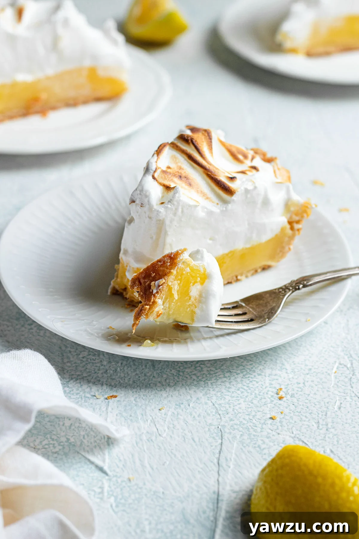 A vibrant slice of lemon meringue pie on a pristine white plate, accompanied by a silver fork, showcasing its golden crust, bright yellow filling, and tall, toasted meringue.