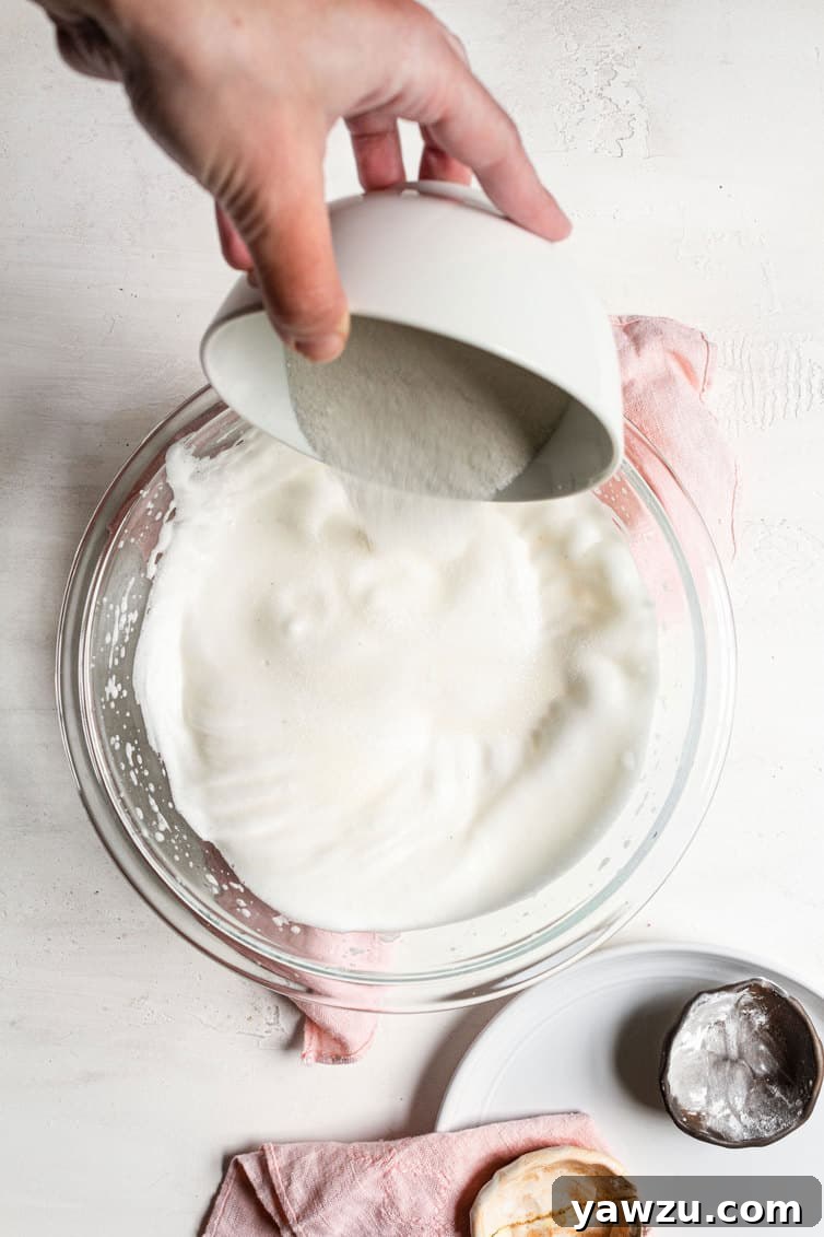 Granulated sugar being slowly added to egg whites already whipped to soft peaks in a mixing bowl.