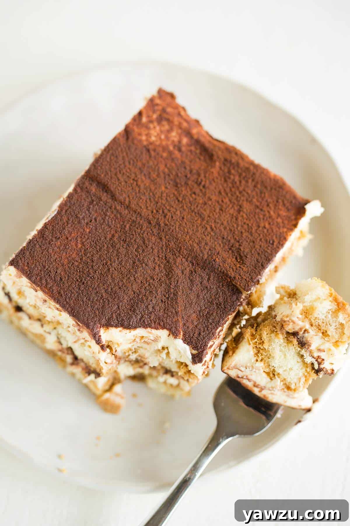 A top down photo of a slice of tiramisu on a white plate with a fork to the right and a bite on the fork.