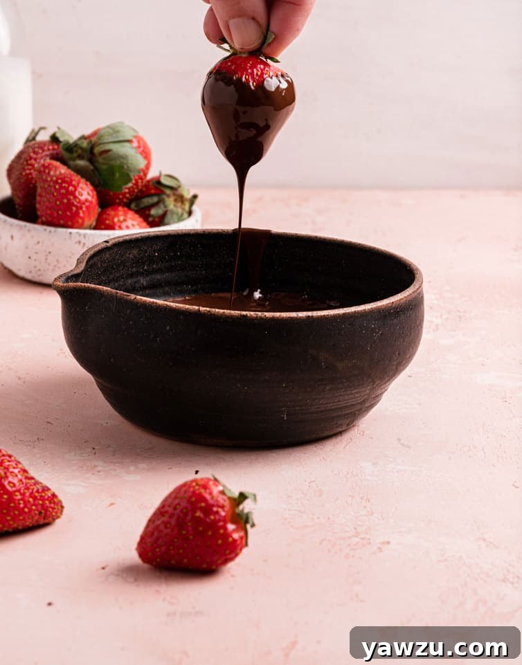 A close-up shot of a hand holding a freshly dipped strawberry, allowing excess melted chocolate to beautifully drizzle back into a brown bowl of rich, liquid chocolate, capturing the elegance of the dipping process.