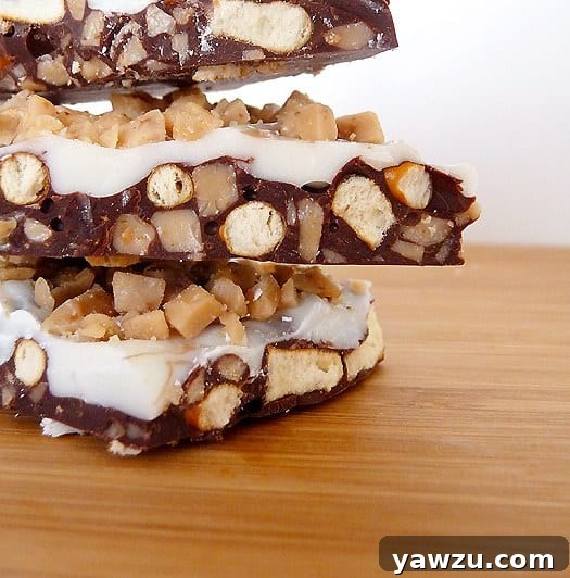 A stack of three pieces of glossy Toffee Pretzel Bark, layered with dark and white chocolate, on a wooden board, showcasing its rich textures.