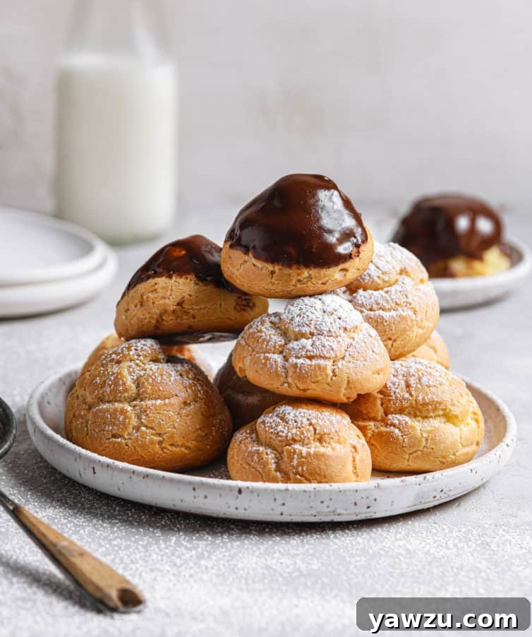 A white plate of chocolate topped cream puffs with powdered sugar cream puffs in a pile with a milk jug in the back.
