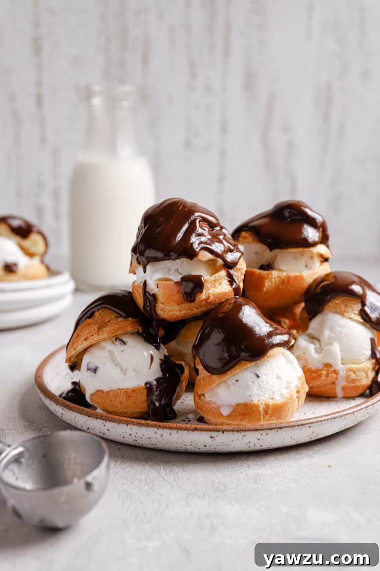 A white plate with a tower of profiteroles topped with chocolate and an ice cream scoop in the front.