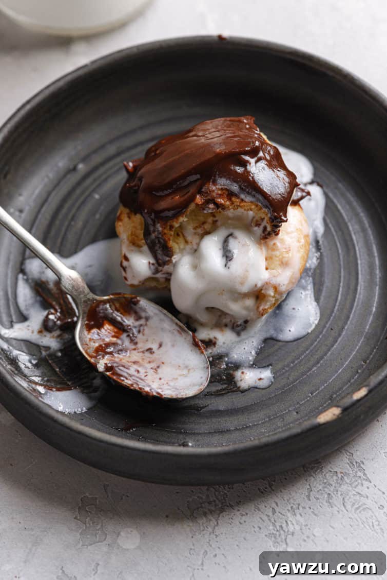 A dark grey plate with an ice cream filled profiterole topped with chocolate sauce and a spoon to the left.
