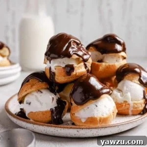 A white plate with a stack of profiteroles topped with melted chocolate sauce and a jar of milk in the back.