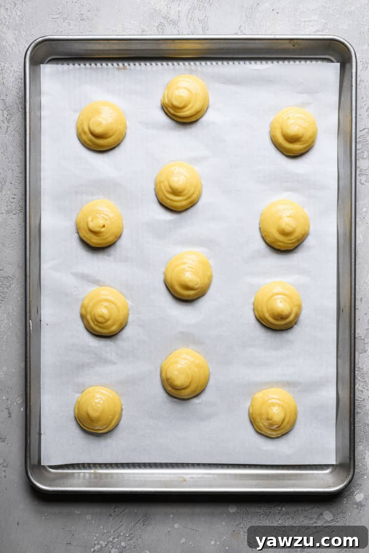 A rimmed baking sheet with a piece of parchment paper with round choux pastry puffs before baking.