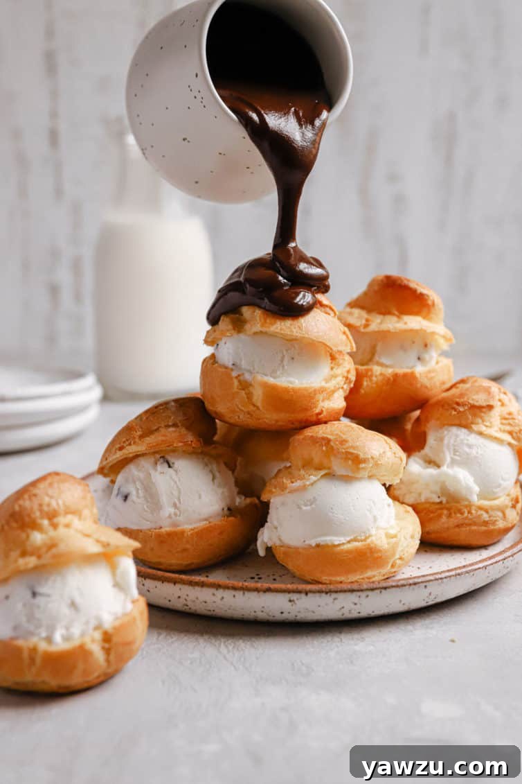 A stack of vanilla ice cream filled profiteroles with a jar of chocolate sauce pouring over the top.