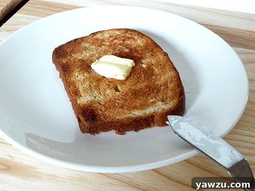 A single slice of honey oatmeal sandwich bread, buttered, on a white plate, ready to eat.