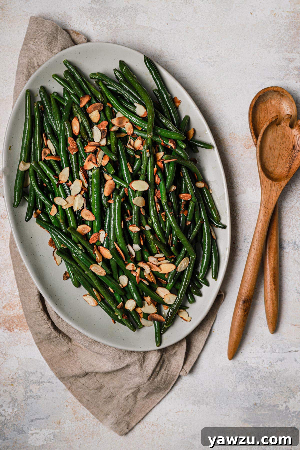 Vibrant green beans generously topped with golden toasted sliced almonds, presented in a white serving dish with wooden serving spoons nearby.