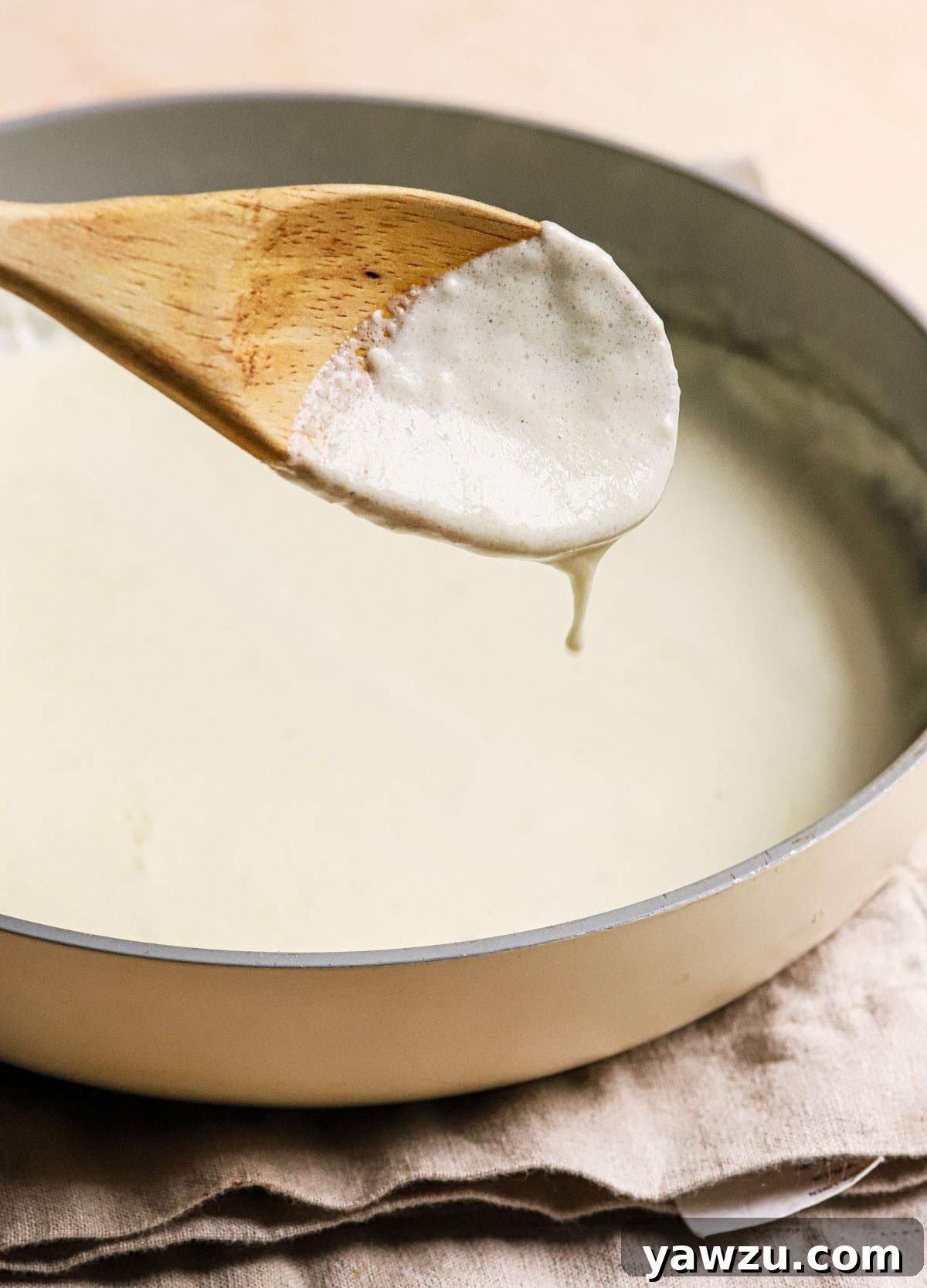 Alfredo sauce dripping off a wooden spoon being held above a saucepan.