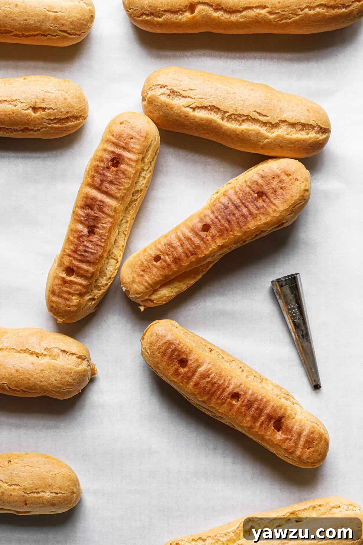 Empty eclair shells on a piece of parchment paper with holes poked in the bottom and a piping tip to the right.