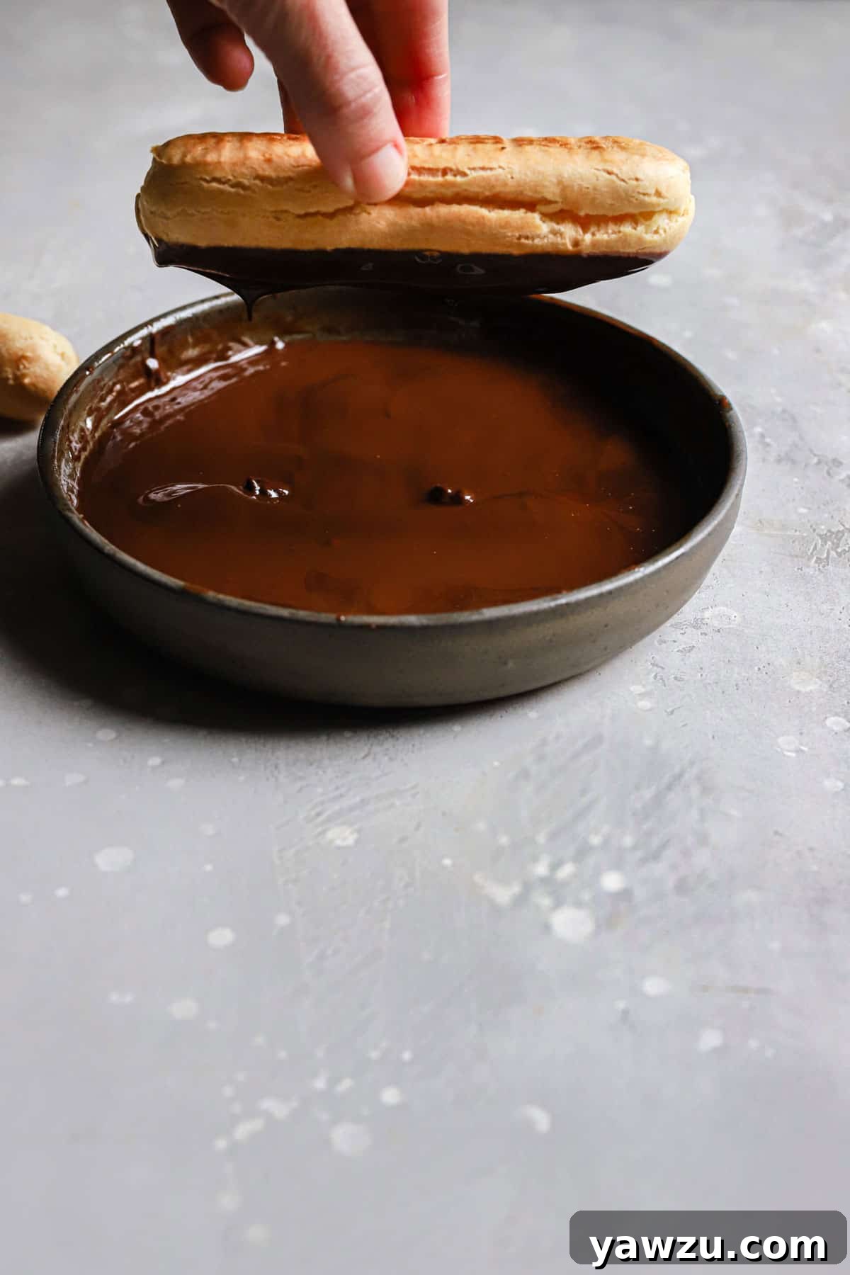 A hand holding an eclair upside down to dip it in the chocolate glaze.