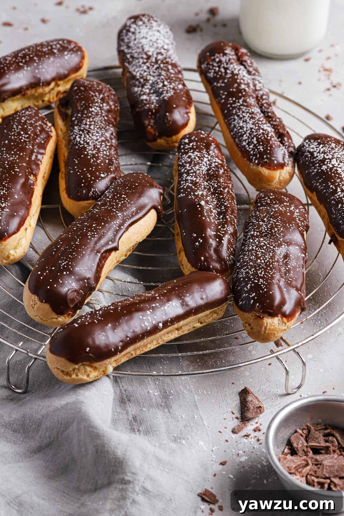 A bowl of cocoa powder in the front right with a wire cooling rack of eclairs topped with chocolate glaze and dusted with powdered sugar.