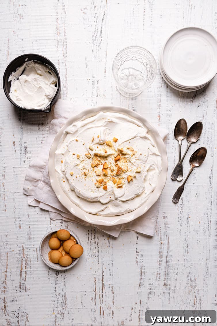 A banana pudding in a pie pan on a towel with spoons on the right a bowl of whipped cream in the top left and a bowl of eggs in the bottom left.