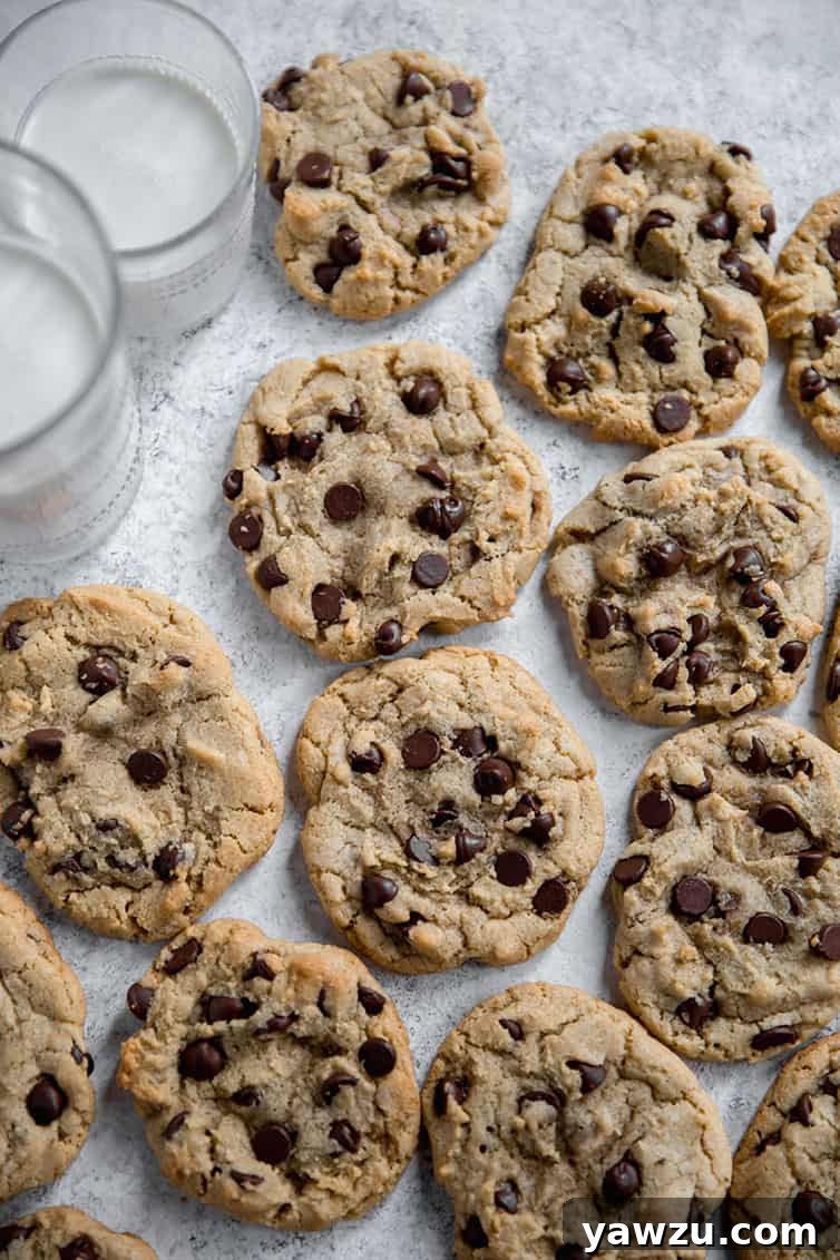 Freshly baked thick and chewy chocolate chip cookies cooling on wax paper, with two glasses of milk beside them.