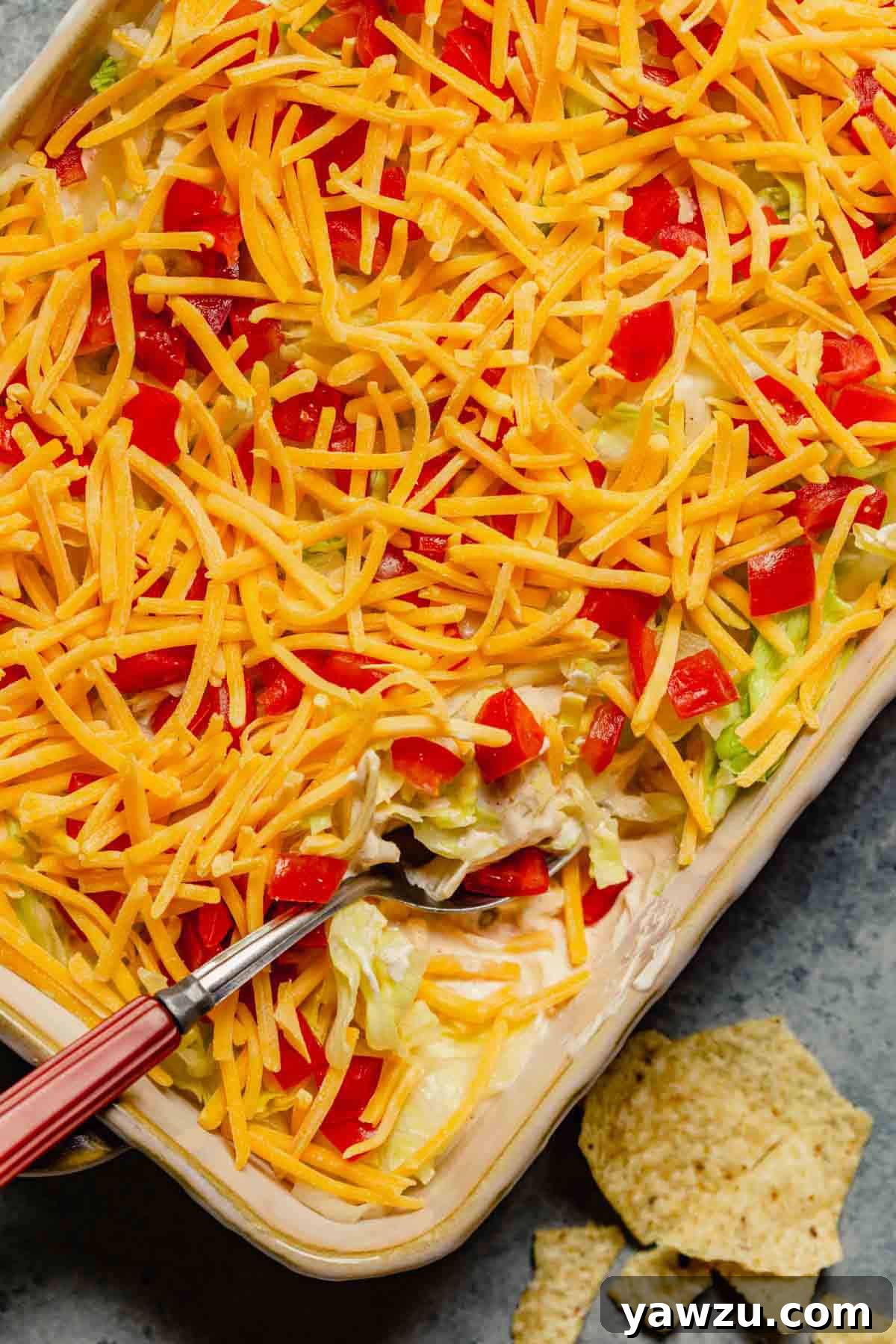 A casserole dish with taco dip and a red handled spoon in the bottom right corner on a counter with a few tortilla chips.