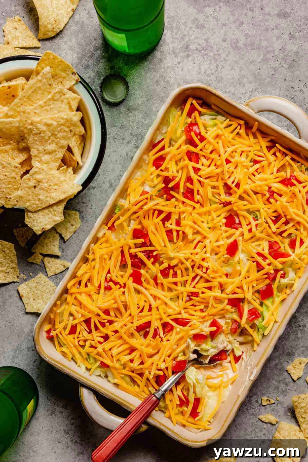 Cheesy no-bake taco dip in a casserole dish with a spoon in the right corner of the dip and a bowl of tortilla chips to the left of the dip.