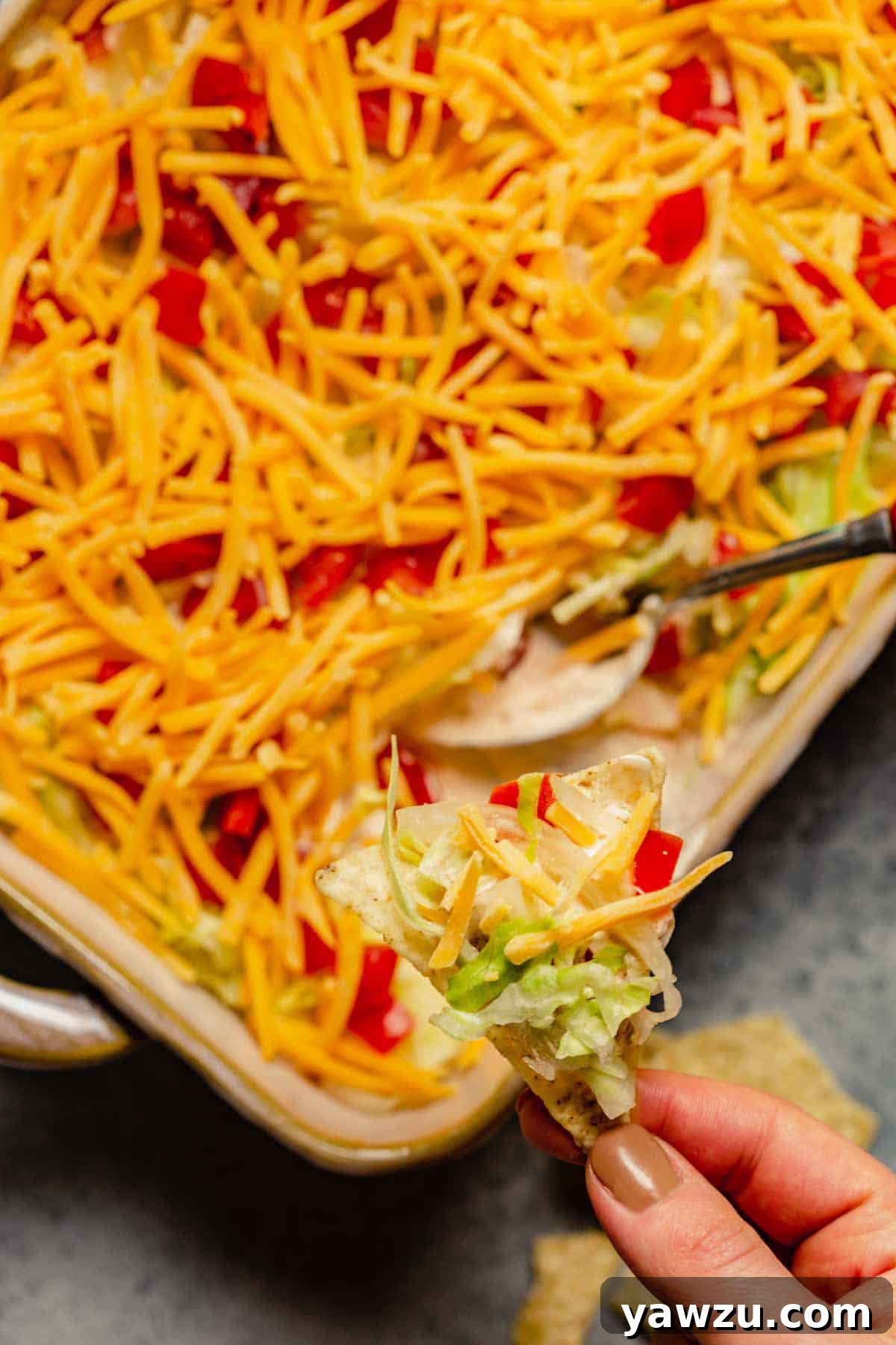 A casserole dish with taco dip and a spoon in the bottom right with a hand holding a tortilla chip above.