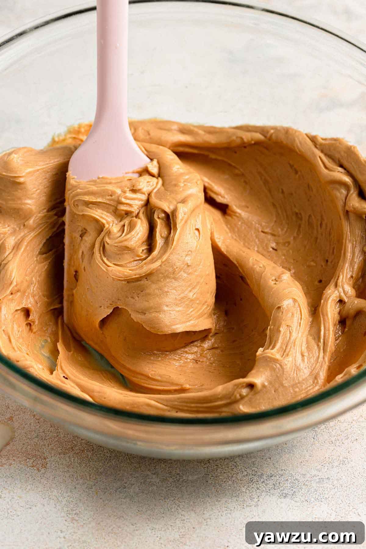 A pink spatula stirring peanut butter frosting in a glass bowl.