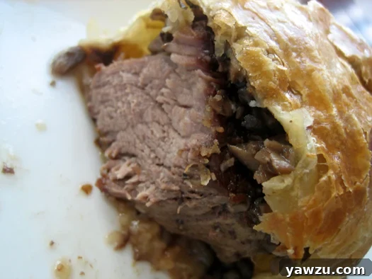 A perfectly cooked individual Beef Wellington, sliced open to reveal a succulent medium-rare filet mignon coated in rich mushroom duxelles and encased in golden, flaky puff pastry.