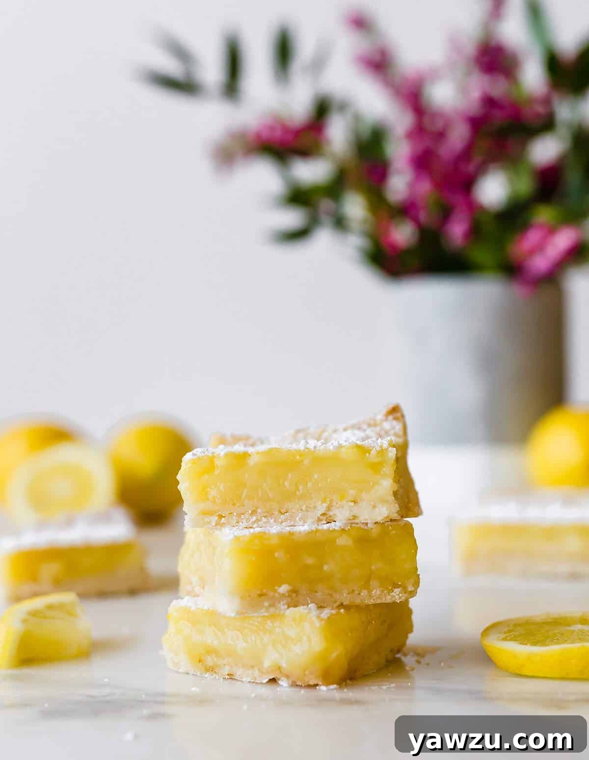 A beautifully arranged stack of three classic lemon bars, garnished with fresh lemon slices and a vase of white flowers in the background, highlighting their bright and inviting appearance.