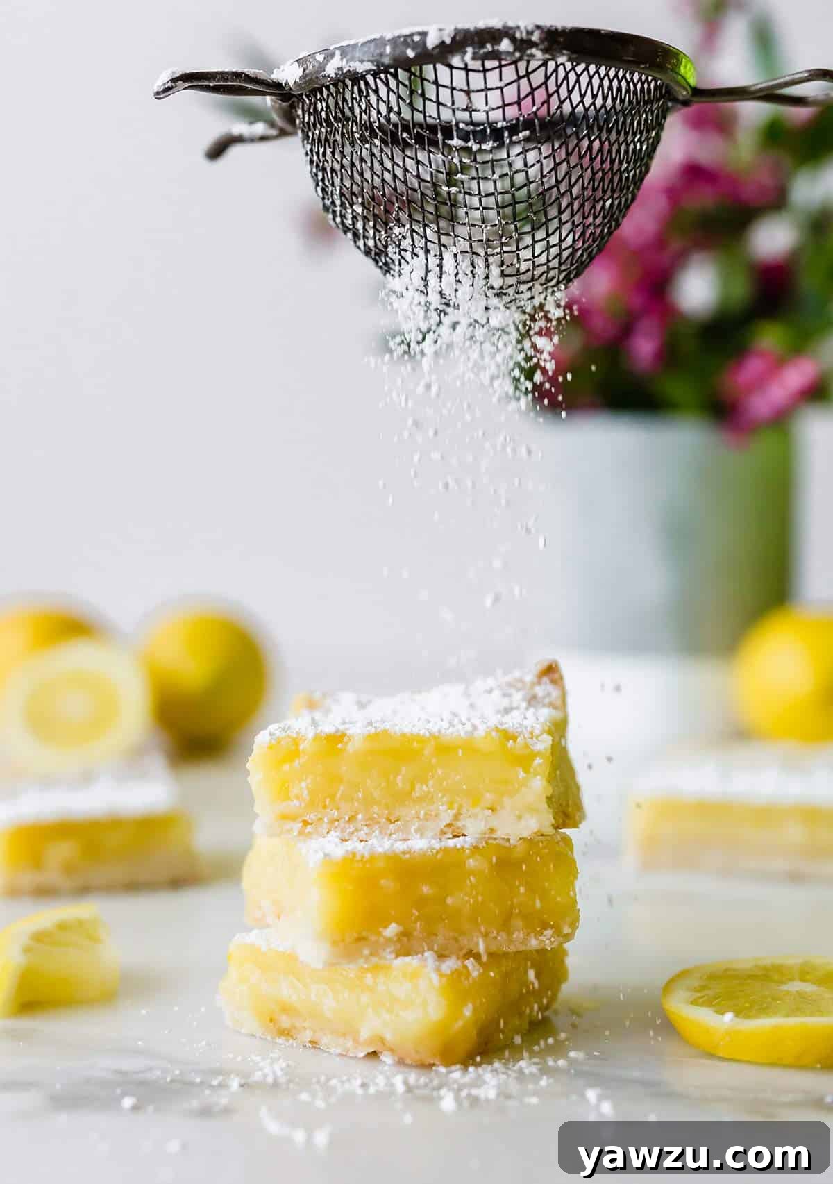A stack of three perfectly golden lemon bars being gracefully sprinkled with a generous dusting of powdered sugar, enhancing their inviting appearance.