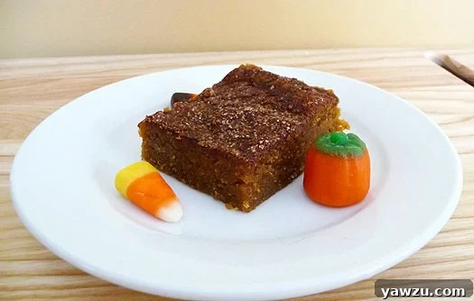 A perfectly portioned square of pumpkin pie bar, generously dusted with a shimmering cinnamon-sugar topping, rests on a pristine white plate, adorned with festive candy corn for a touch of autumn charm. The bar's rich, golden-brown hue and moist, dense texture are inviting, promising a comforting taste of fall.