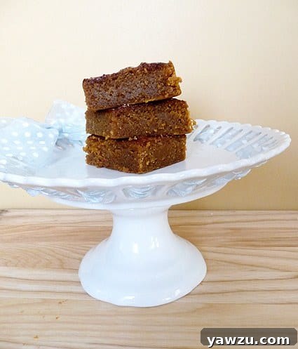 A charming stack of three delectable pumpkin pie bars, each glistening with a cinnamon-sugar topping, thoughtfully arranged on a quaint white dessert stand. The image highlights the rich, soft interior texture and the appealing thickness of each bar, ready for a comforting bite.