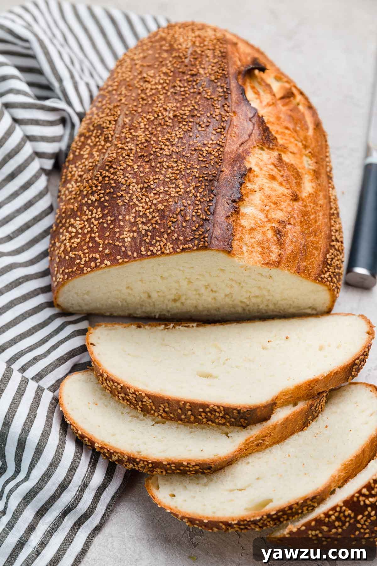 Loaf of Italian bread with sesame seeds on top, with four slices sliced off and laying in front of loaf with napkin and knife to the side.